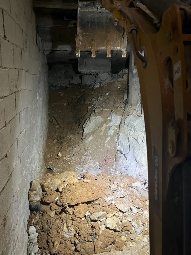 חפירת מרתף מתחת לבית קיים: כף מיני מחפרון בחלל צר בין קיר בלוקים לסלע חשוף