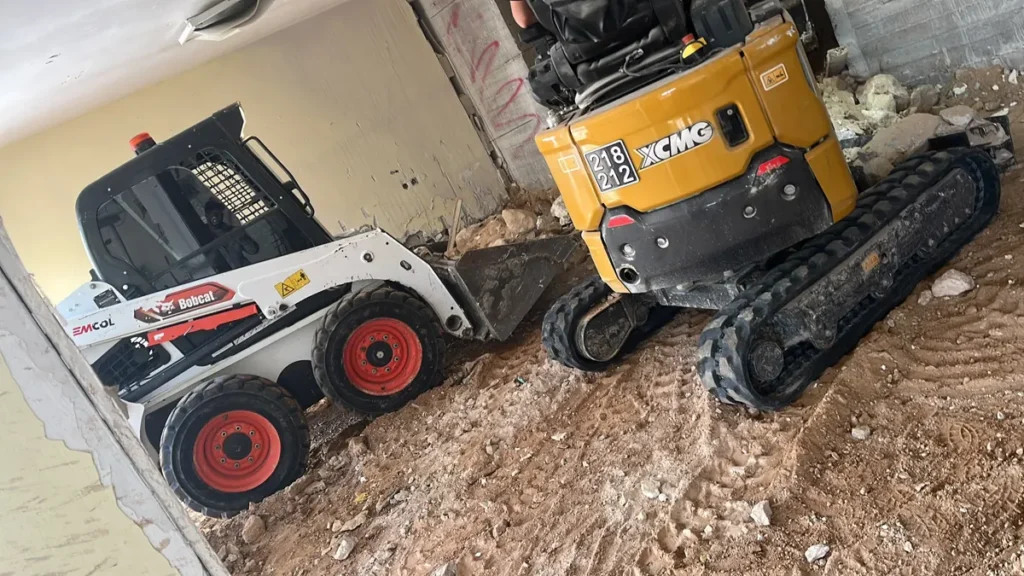 בובקט עם כף ומיני מחפרון זחלי זה לצד זה בתוך מבנה, על רצפת עפר חפורה