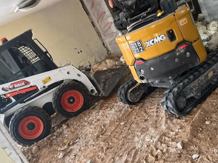 בובקט עם כף ומיני מחפרון זחלי זה לצד זה בתוך מבנה, על רצפת עפר חפורה