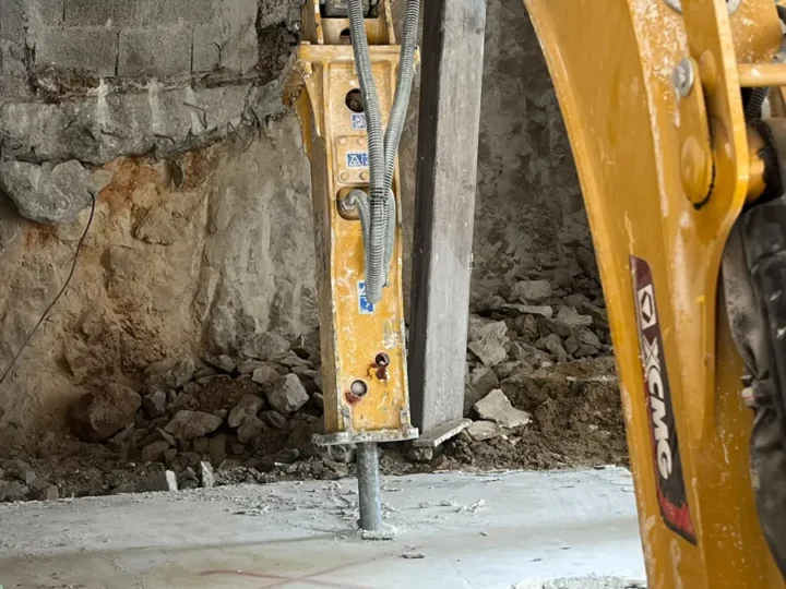 פטיש הידראולי על מיני מחפרון שובר רצפת בטון בתוך מבנה, ברקע דופן סלע וקיר בלוקים