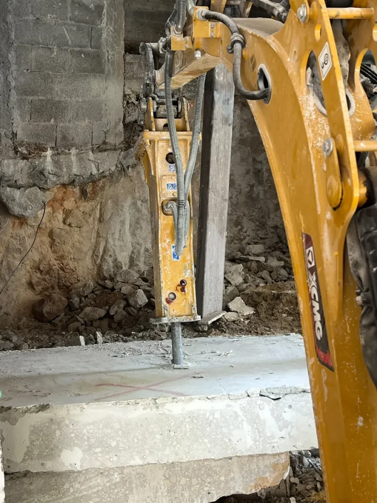פטיש הידראולי על מיני מחפרון שובר רצפת בטון בתוך מבנה, ברקע דופן סלע וקיר בלוקים