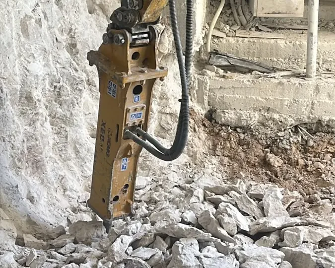 פטיש הידראולי על מיני מחפרון חוצב בסלע גיר לבן צמוד ליסודות מבנה קיים