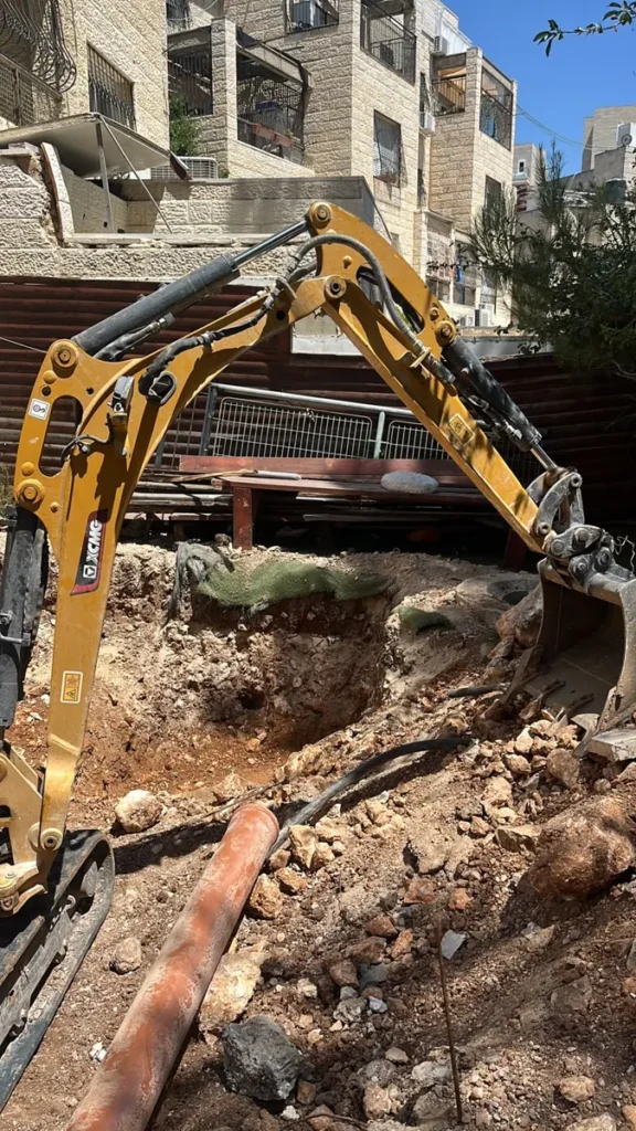 מיני מחפרון עם כף חופר בור בחצר צרה בין בנייני אבן, לצד צינור ביוב כתום
