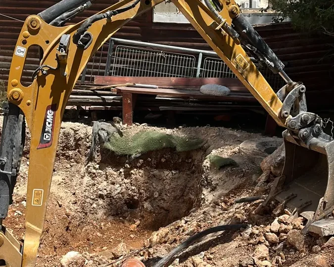 מיני מחפרון עם כף חופר בור בחצר צרה בין בנייני אבן, לצד צינור ביוב כתום