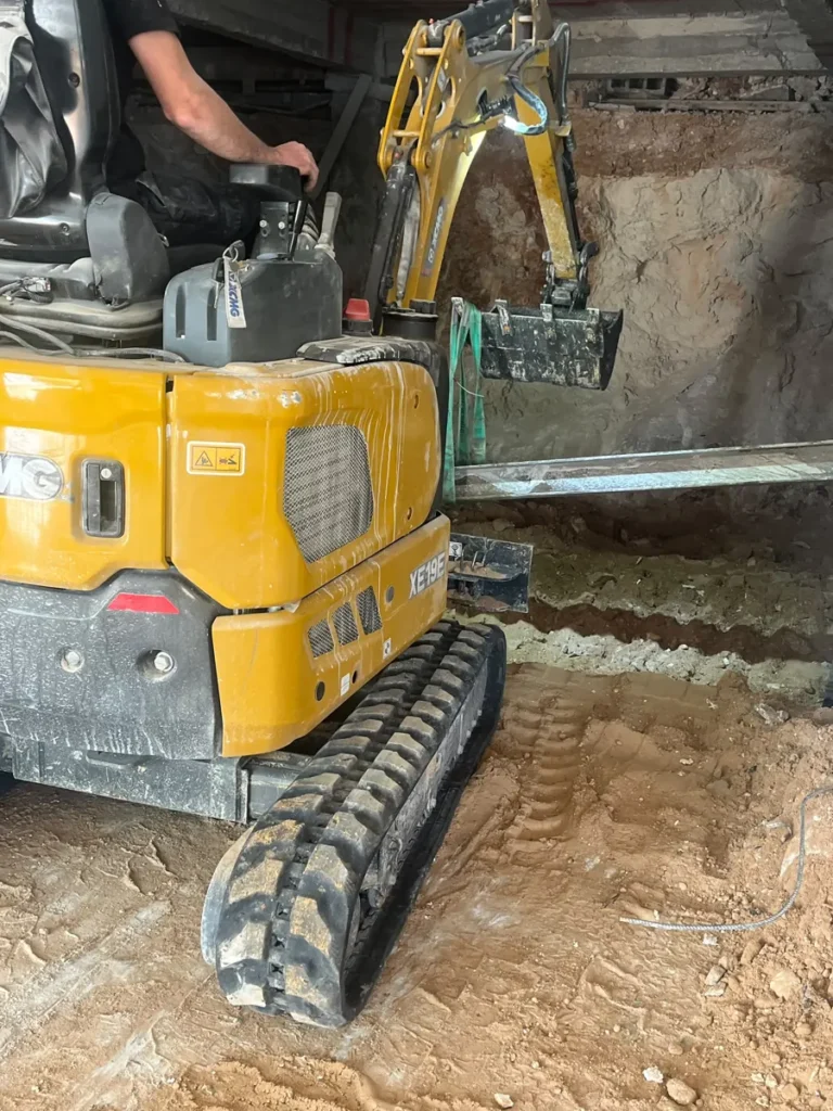 מיני מחפרון חופר מרתף בתוך מבנה קיים מתחת לתקרה, ליד קורת פלדה תומכת