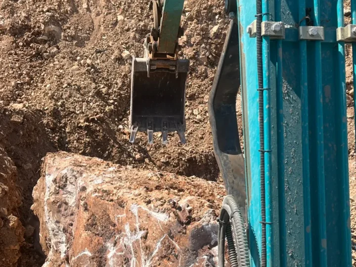 מיני מחפרון חופר באדמה אדמדמה סלעית, ולידו סלע גדול שנחשף בחפירה