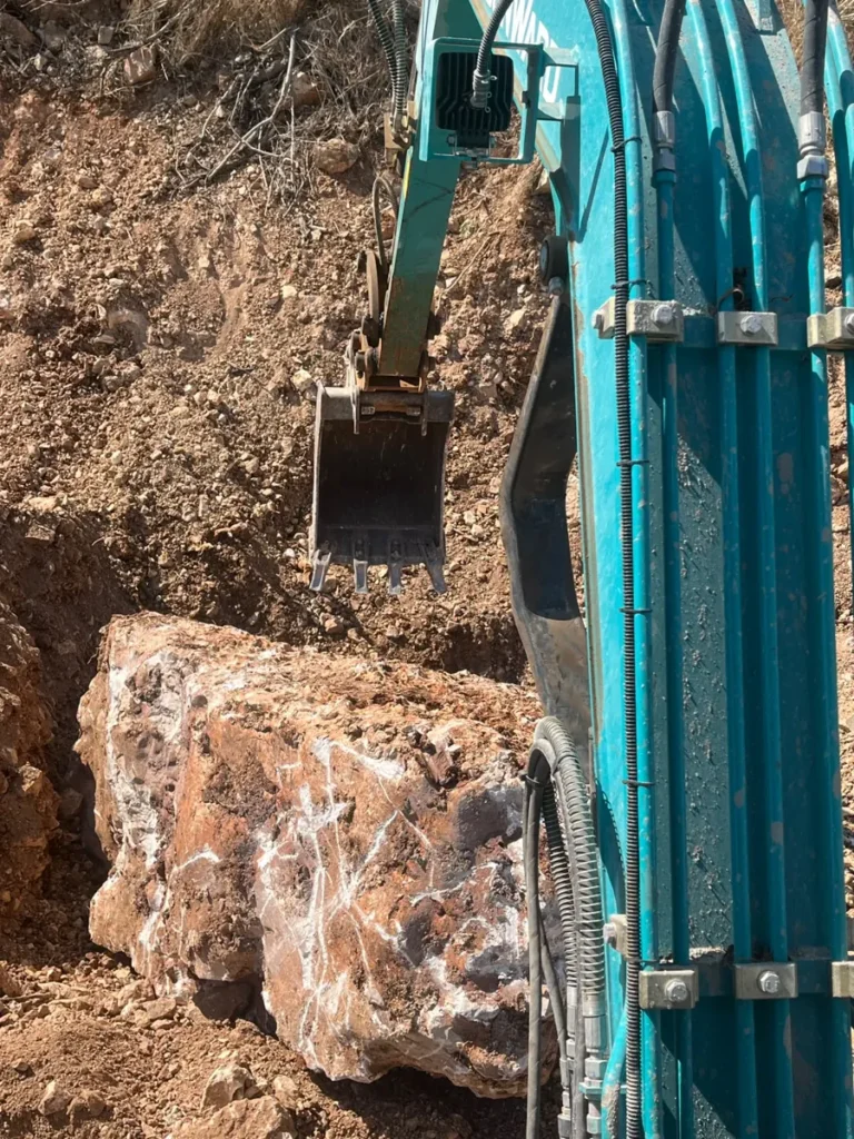 מיני מחפרון חופר באדמה אדמדמה סלעית, ולידו סלע גדול שנחשף בחפירה