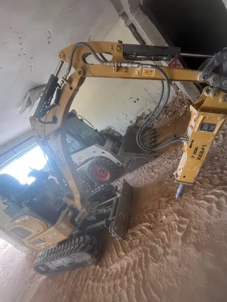 מיני מחפרון עם פטיש הידראולי ובובקט עובדים יחד בתוך חדר בקומת קרקע