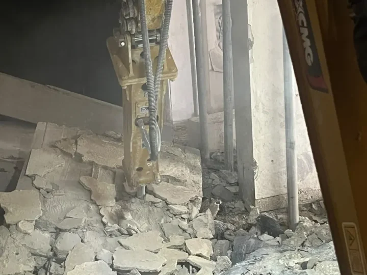 פטיש הידראולי על מיני מחפרון שובר רצפת בטון בתוך מבנה, בין תמוכות פלדה