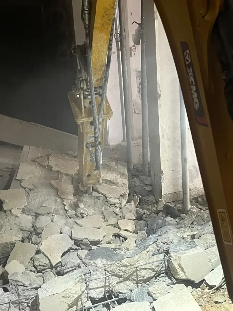 פטיש הידראולי על מיני מחפרון שובר רצפת בטון בתוך מבנה, בין תמוכות פלדה