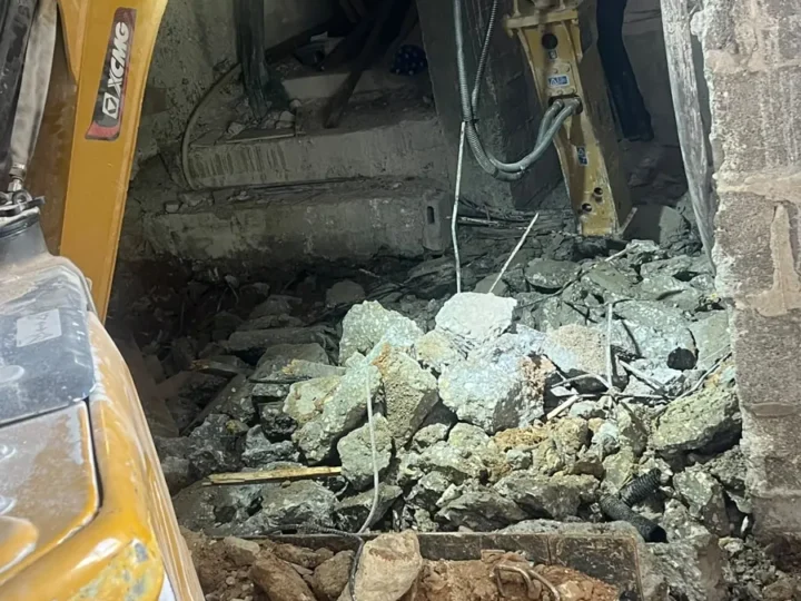 מיני מחפרון עם פטיש הידראולי שובר מדרגות בטון בתוך מבנה, ושברי בטון וברזל על הרצפה