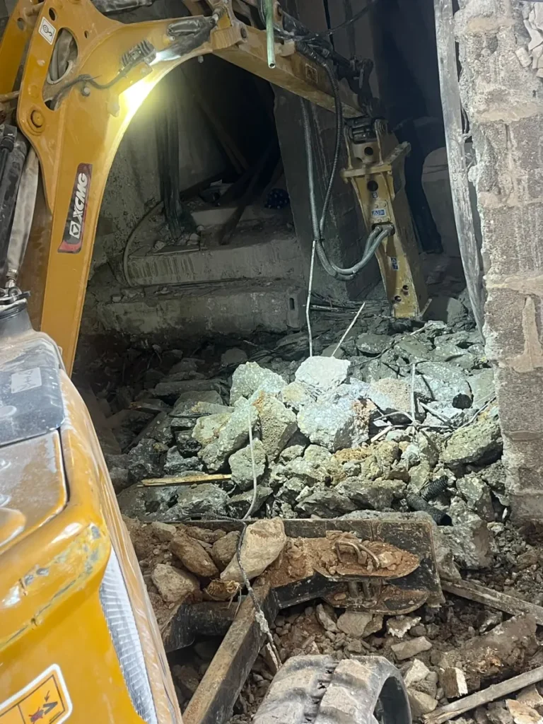 מיני מחפרון עם פטיש הידראולי שובר מדרגות בטון בתוך מבנה, ושברי בטון וברזל על הרצפה