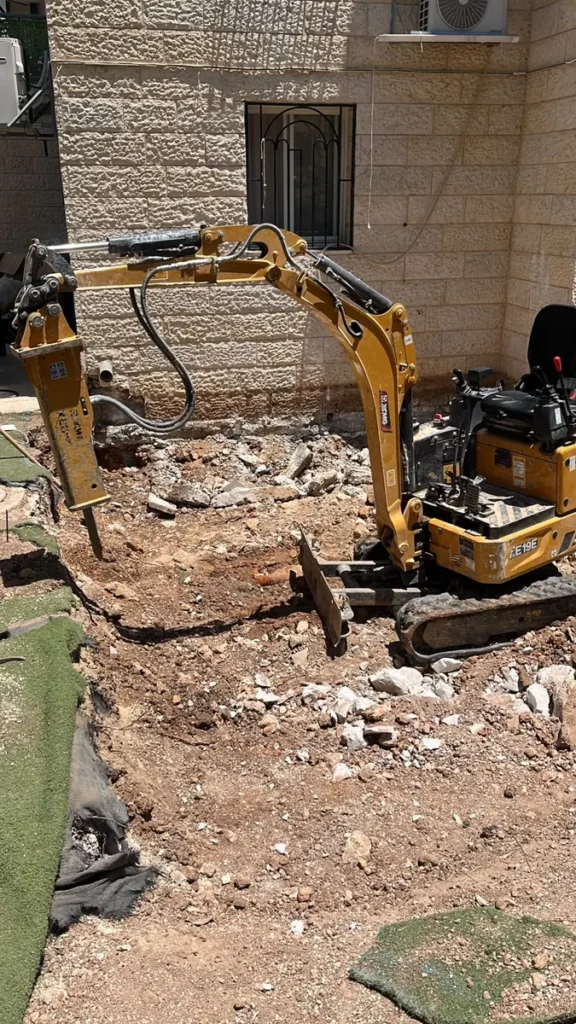 מיני מחפרון עם פטיש הידראולי חוצב סלע בחצר צמודה לקיר בית אבן