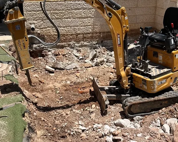 מיני מחפרון עם פטיש הידראולי חוצב סלע בחצר צמודה לקיר בית אבן