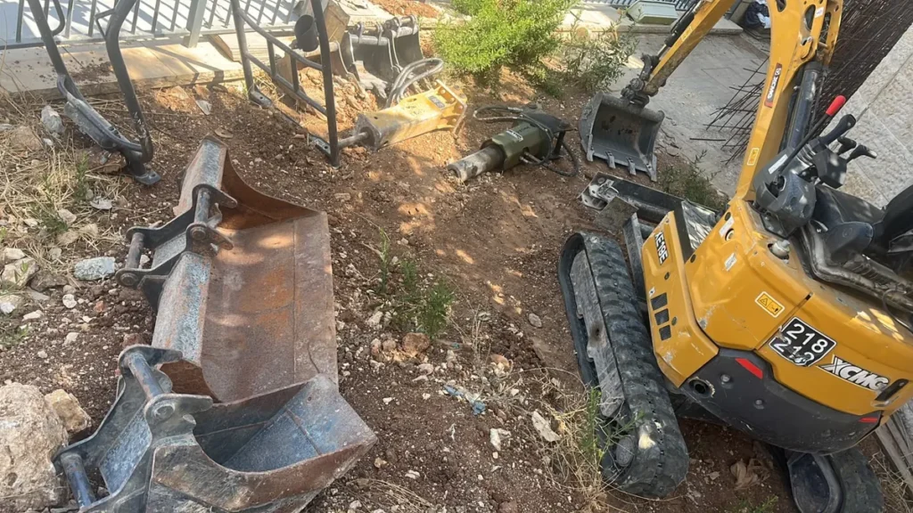 מיני מחפרון זחלי לצד שני פטישים הידראוליים וכפות חפירה בגדלים שונים בחצר עבודה