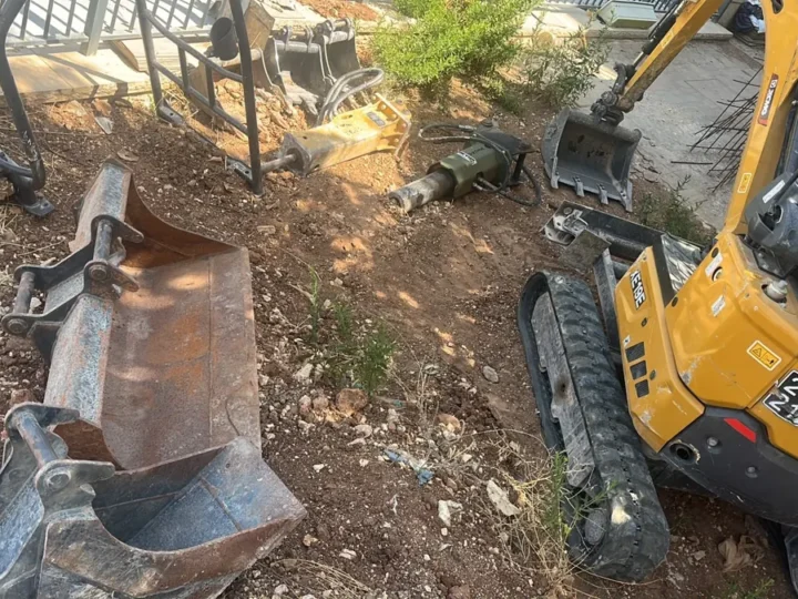 מיני מחפרון זחלי לצד שני פטישים הידראוליים וכפות חפירה בגדלים שונים בחצר עבודה
