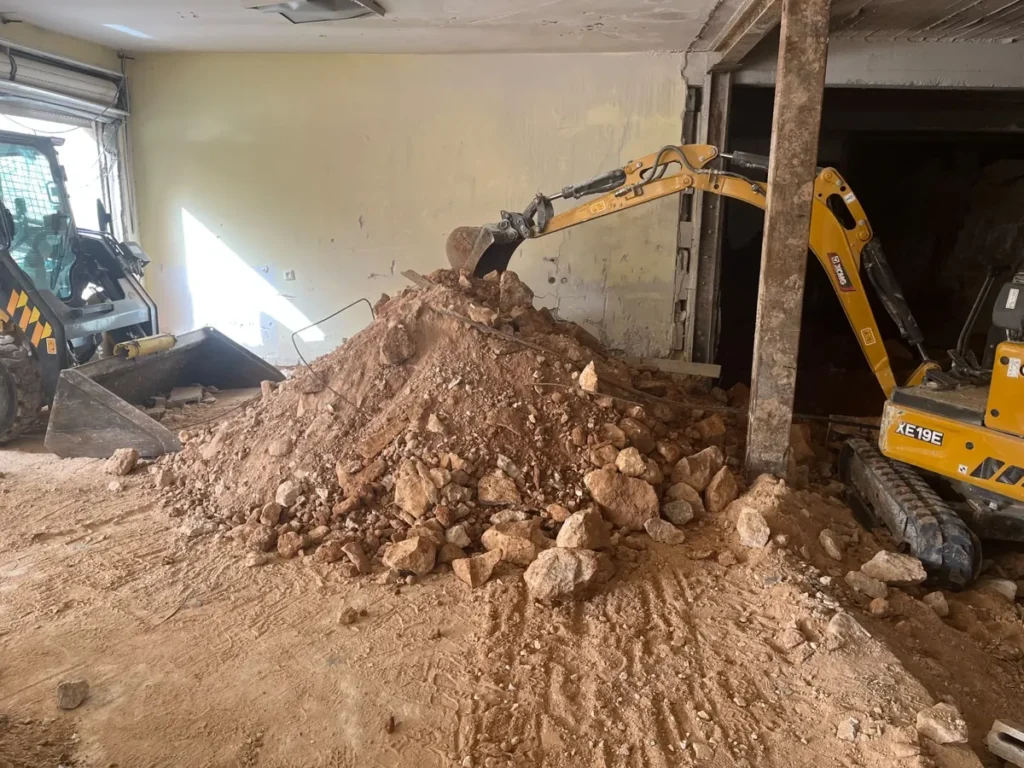מיני מחפרון מערים סלעים ועפר בתוך מבנה קיים ליד עמוד תמיכה, ובובקט ממתין לפינוי