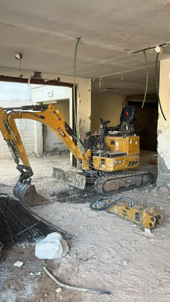 מיני מחפרון בתוך קומת קרקע של בית קיים, ולידו פטיש הידראולי מנותק המוכן להחלפה