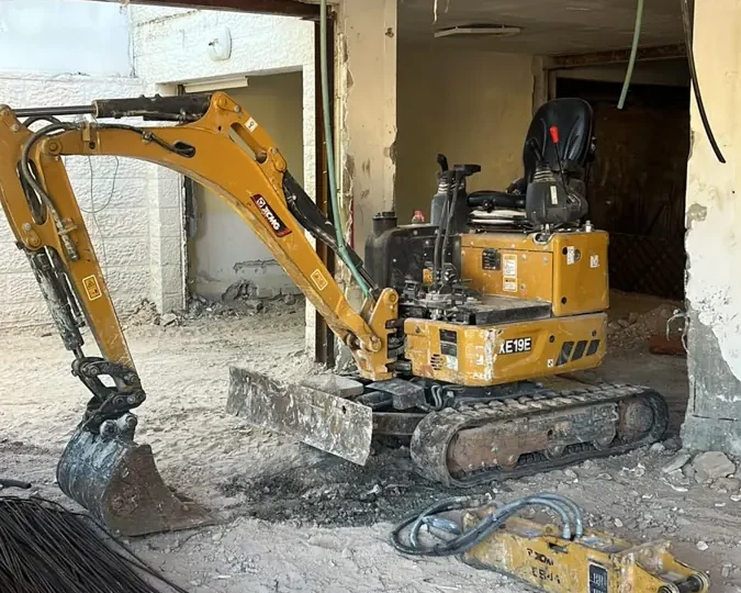 מיני מחפרון בתוך קומת קרקע של בית קיים, ולידו פטיש הידראולי מנותק המוכן להחלפה