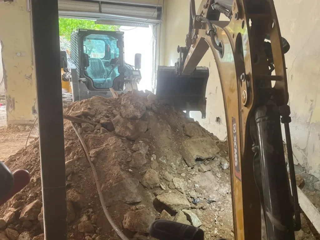 מיני מחפרון מעמיס ערמת עפר ואבנים בתוך מבנה, ובובקט ממתין בפתח לפינוי החומר