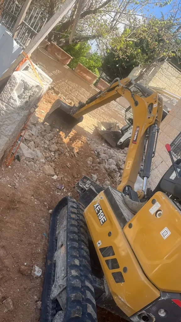 מיני מחפרון קטן ובובקט עובדים בחצר צרה בין קירות אבן, בחפירה ופינוי של אדמה ושברי אבן