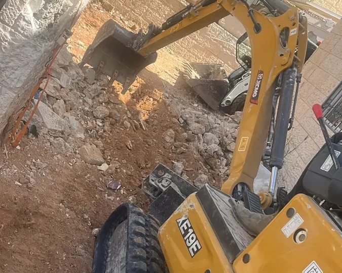 מיני מחפרון קטן ובובקט עובדים בחצר צרה בין קירות אבן, בחפירה ופינוי של אדמה ושברי אבן