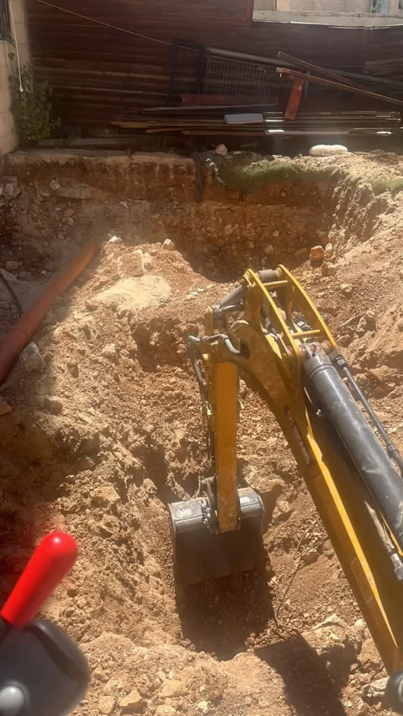 מבט ממושב המפעיל: מיני מחפרון חופר בור בחצר בקרקע אדומה ואבנים
