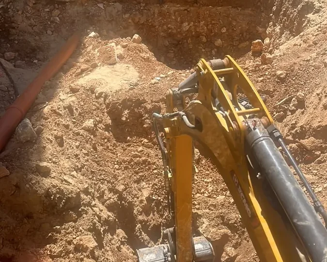 מבט ממושב המפעיל: מיני מחפרון חופר בור בחצר בקרקע אדומה ואבנים