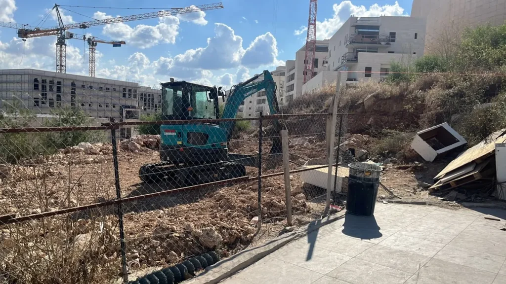 מחפר זחלי עם תא נהג מכשיר מגרש סלעי, ברקע מנופי בנייה ובניינים חדשים