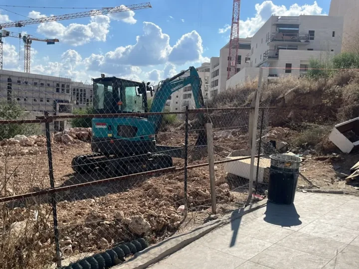 מחפר זחלי עם תא נהג מכשיר מגרש סלעי, ברקע מנופי בנייה ובניינים חדשים